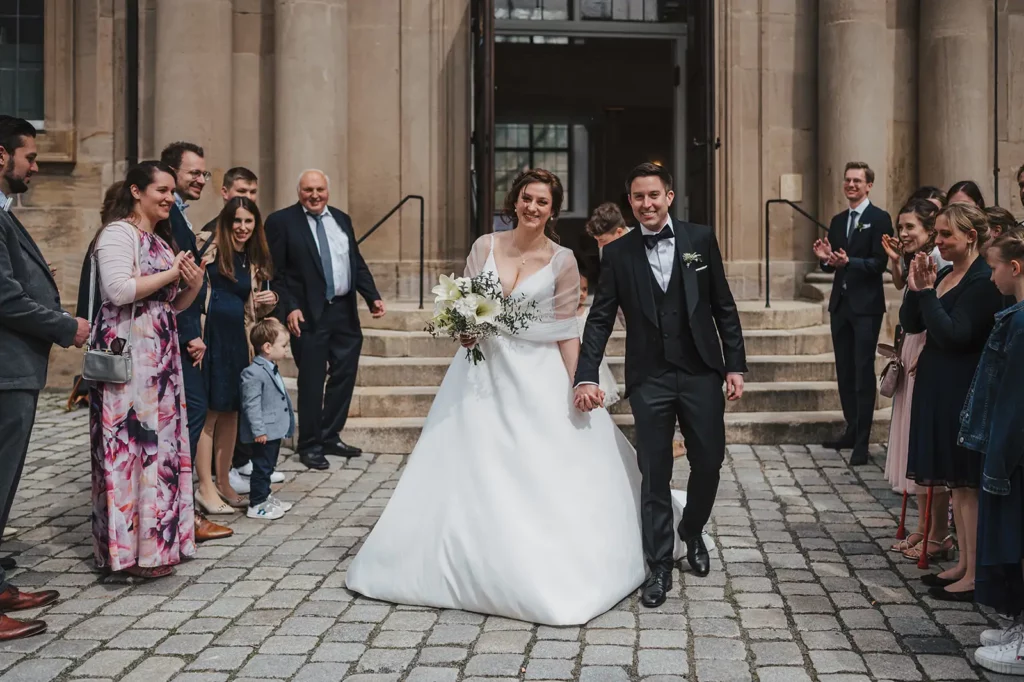 Brautpaar verlässt die Kirche bei der Hochzeit im Liebesbier Bayreuth, begleitet von applaudierenden Gästen.