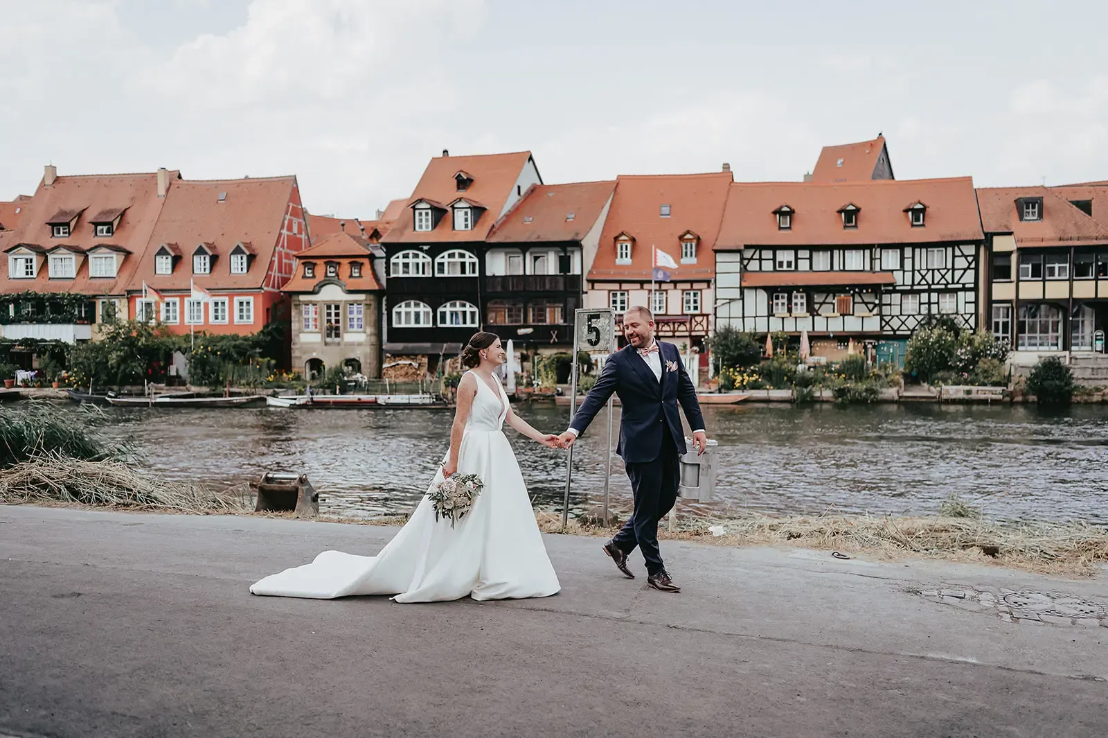 Lena und Günther spazieren am Flussufer in Bamberg entlang. Im Hintergrund sind Fachwerkhäuser und historische Gebäude zu sehen.