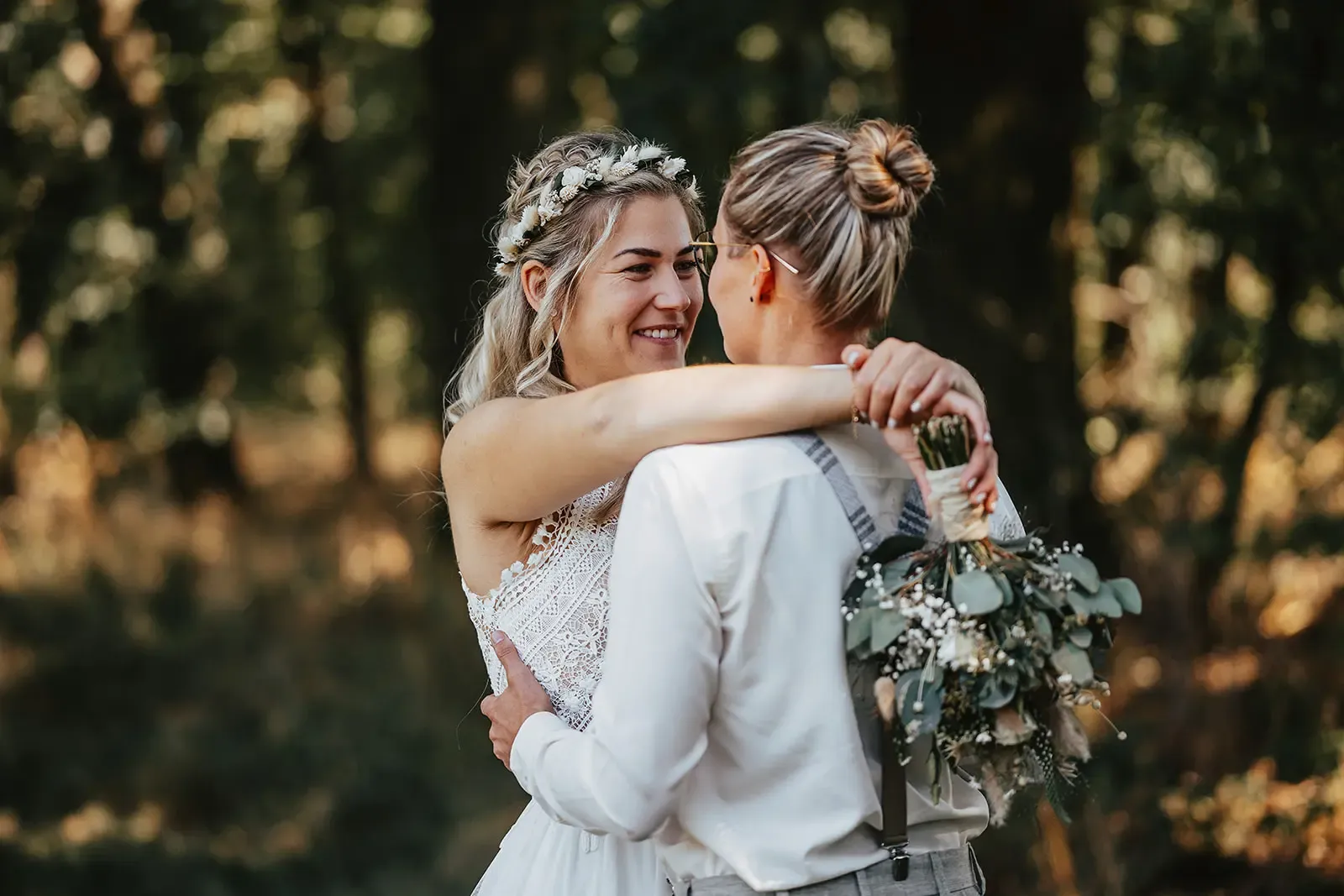 Das Hochzeitspaar umarmt sich liebevoll im Wald der Osterburg Weida, die Braut im weißen Kleid und die Partnerin im grauen Anzug mit Hosenträgern.